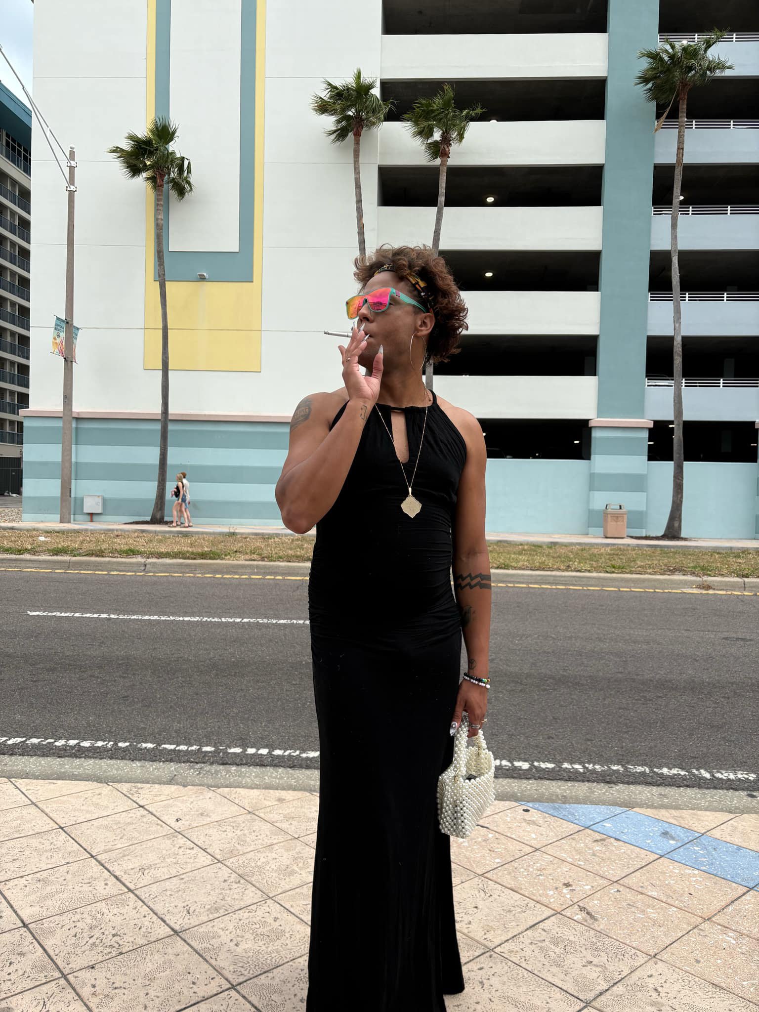 a mixed race browned skin trans-fem wearing a black dress and smoking a cigarettes with a parking structure and palms trees in the background